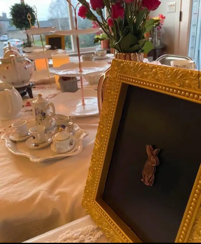 A table topped with a fancy gold frame picture of a bunny, flowers, and a teacup set