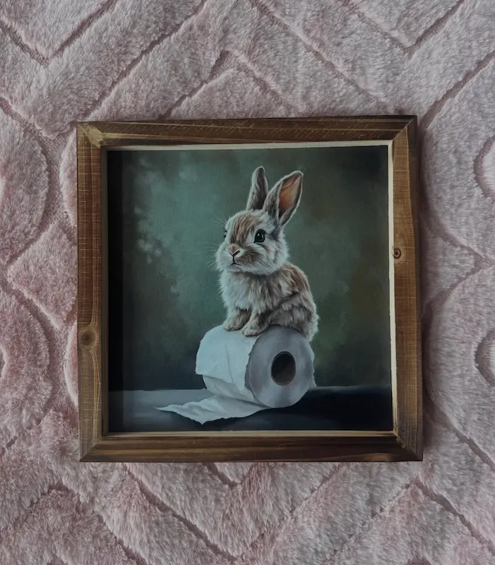 Framed painting of a brown bunny sitting atop a roll of toilet paper
